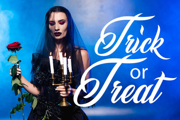 woman in black dress and veil holding red rose and candles near trick or treat lettering on blue with smoke