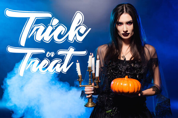 pale woman with scary makeup holding pumpkin and burning candles near trick or treat lettering on blue with smoke