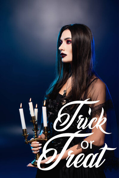 woman in black dress and veil holding burning candles near trick or treat lettering on blue 