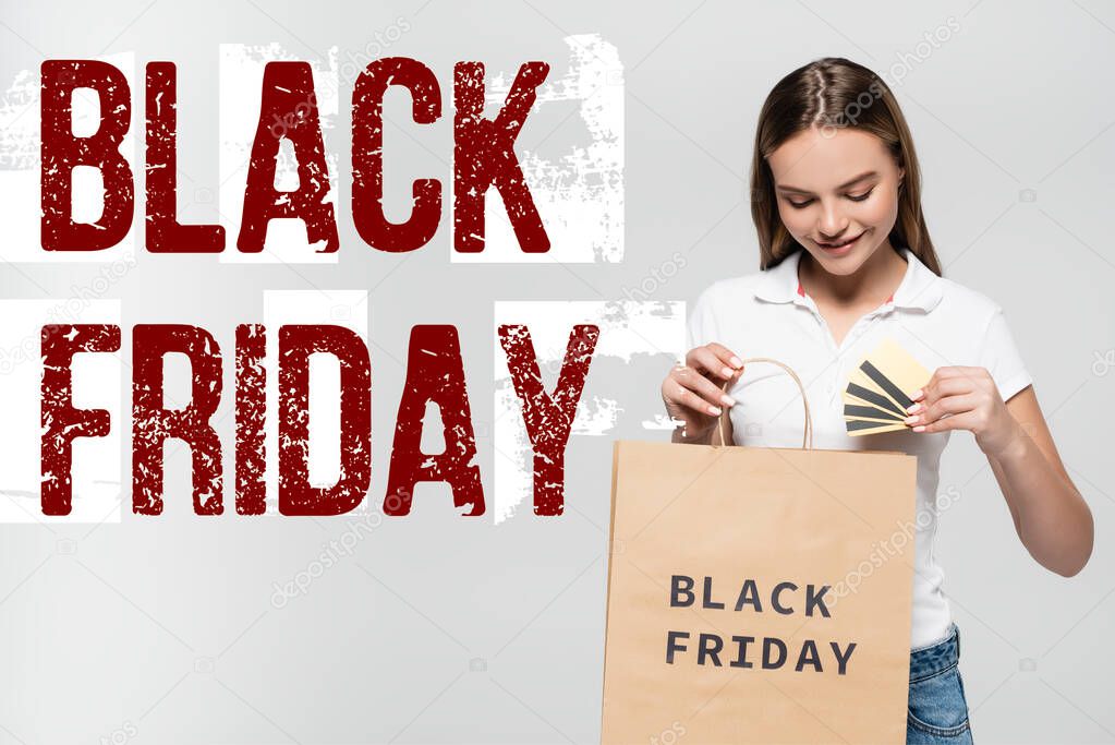 joyful woman holding credit cards and looking at shopping bag near black friday lettering on grey