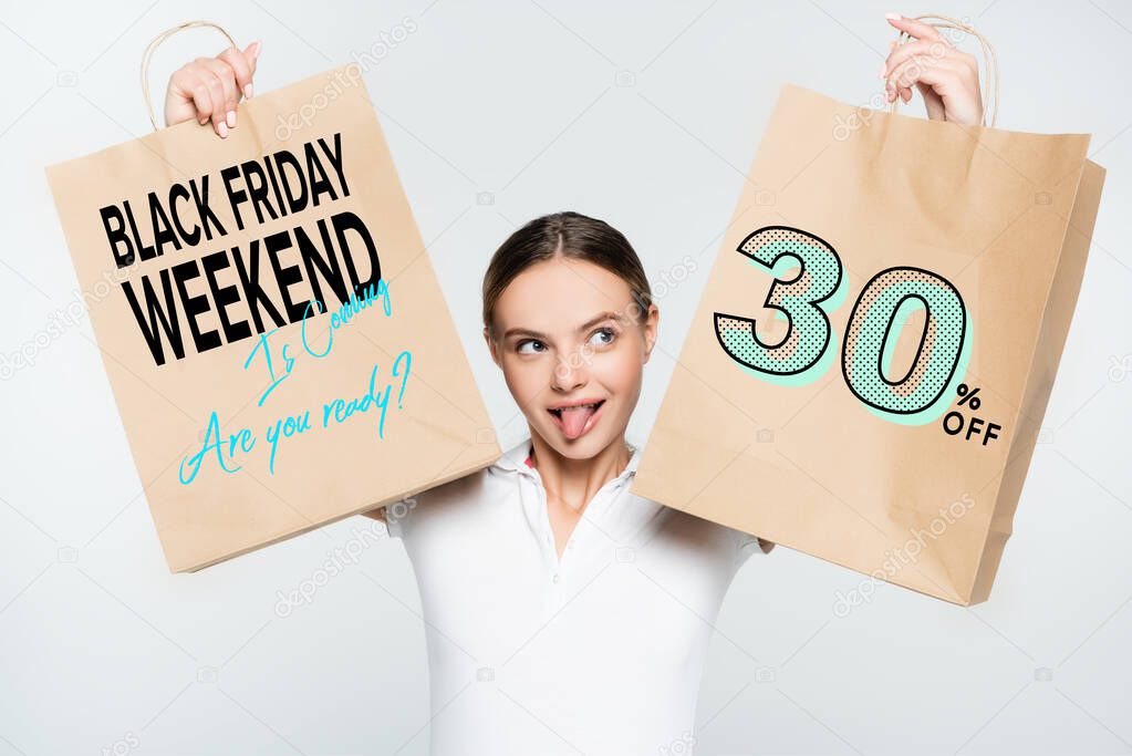 young woman sticking out tongue while holding shopping bags with black friday weekend lettering isolated on white