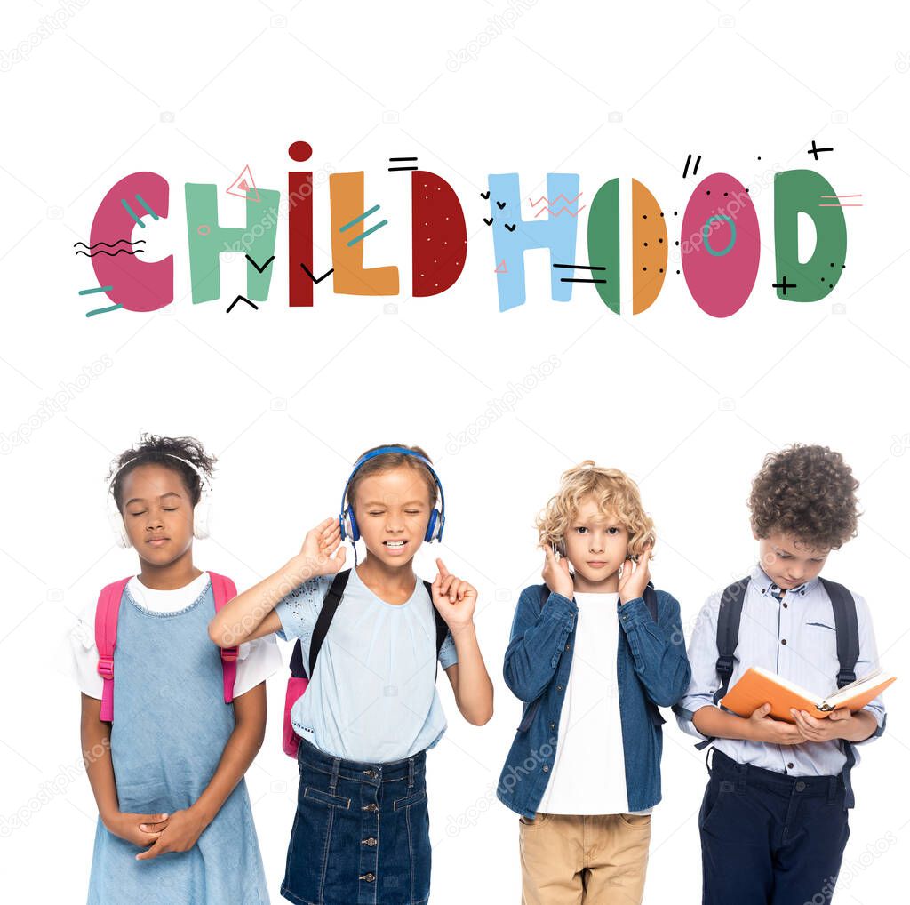 multicultural schoolkids listening music in wireless headphones near curly boy reading book and childhood lettering on white 