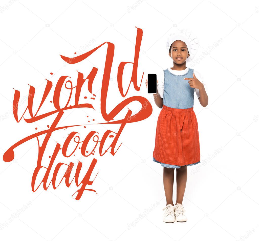 african american kid dressed in costume of chef pointing with finger at smartphone with blank screen near world food day lettering on white 