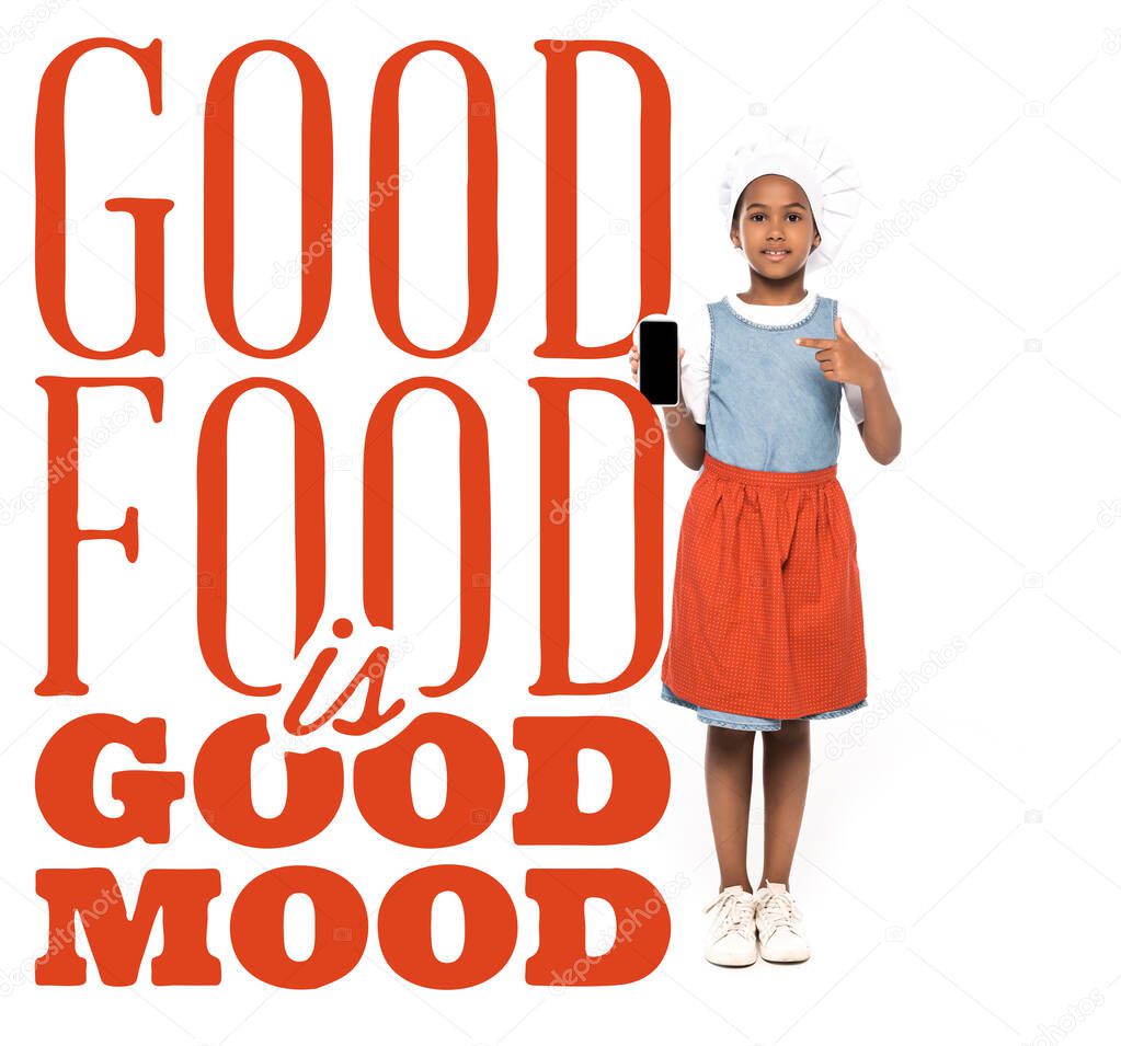 african american kid dressed in costume of chef pointing with finger at smartphone near good food is good mood lettering on white 
