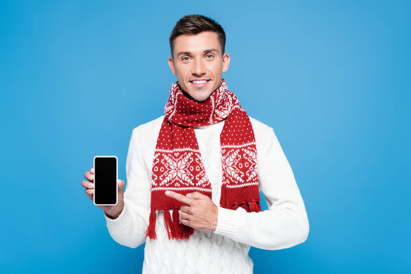 Smiling young adult man, wearing sweater and scarf, showing and pointing with finger at smartphone isolated on blue