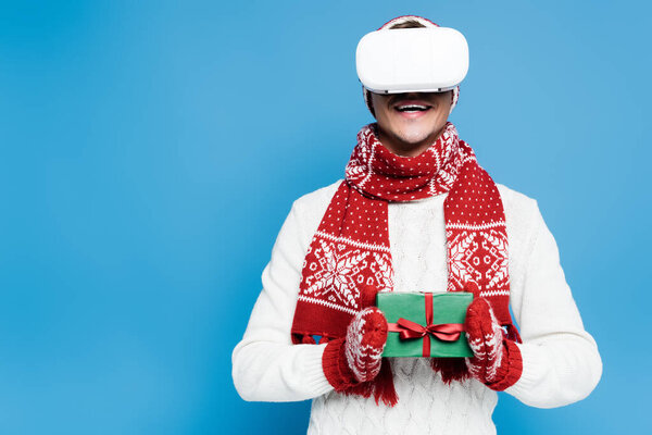 Smiling man in warm clothing, using vr headset and holding small gift box isolated on blue
