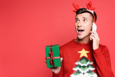 Çam ağacından süveter giymiş heyecanlı bir adam akıllı telefondan konuşuyor ve kırmızı üzerine izole edilmiş bir hediye kutusu tutuyor.
