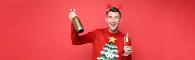 Cheerful man in Christmas sweater holding glass and bottle of champagne isolated on red, banner clipart