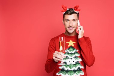 Kırmızı arka planda bir bardak şampanyayla gülümseyen adam akıllı telefondan konuşuyor.