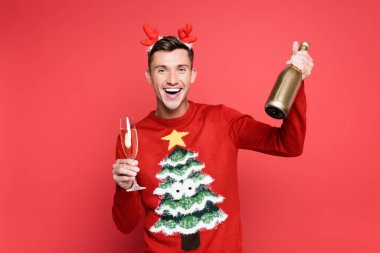 Cheerful man in christmas sweater and headband holding bottle and glass of champagne on red background clipart