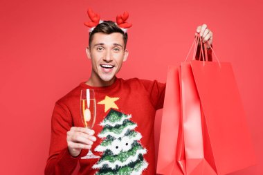 Smiling man in chrismas sweater holding glass of champagne and shopping bags isolated on red clipart