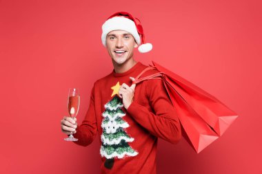 Smiling man in santa hat holding shopping bags and glass of champagne on red background clipart