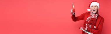 Positive man in santa hat and mittens pointing at flute of champagne on red background, banner clipart