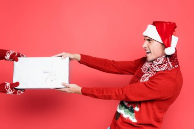 Noel Baba şapkalı ve eşarplı heyecanlı adam kırmızı arka planda hediye kutusunu çekiyor.