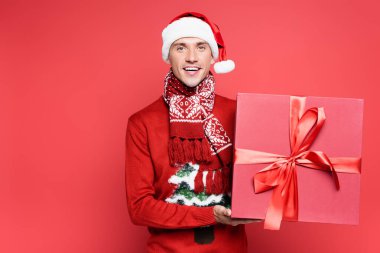 Süveter ve Noel Baba şapkalı neşeli adam kırmızı arka planda hediye kutusu tutuyor.