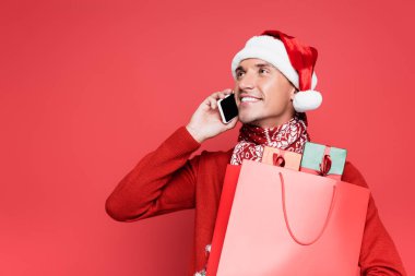 Noel Baba şapkalı gülümseyen adam akıllı telefonla konuşuyor ve elinde kırmızı arka planda hediyeler olan bir alışveriş çantası tutuyor.