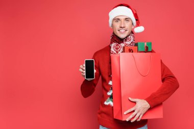 Noel Baba şapkalı genç adam akıllı telefon gösteriyor ve elinde kırmızı arka planda hediye paketleri var.