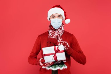 Noel Baba şapkalı, tıbbi maskeli ve eldiven takan genç adam kırmızı kutuda izole edilmiş. 