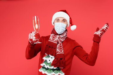 Man in medical mask and santa hat holding glass of champagne isolated on red  clipart