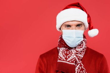Noel Baba şapkalı ve tıbbi maskeli genç adam kırmızı kameraya bakıyor. 