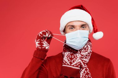 Noel Baba şapkalı ve eldivenli genç adam kırmızı üzerine izole edilmiş tıbbi maske tutuyor. 
