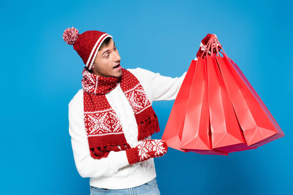 Surprised young adult man in warm clothing holding and looking at red paper bags isolated on blue
