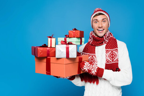 Happy man in knitted mittens and hat holding bunch of gift boxes isolated on blue