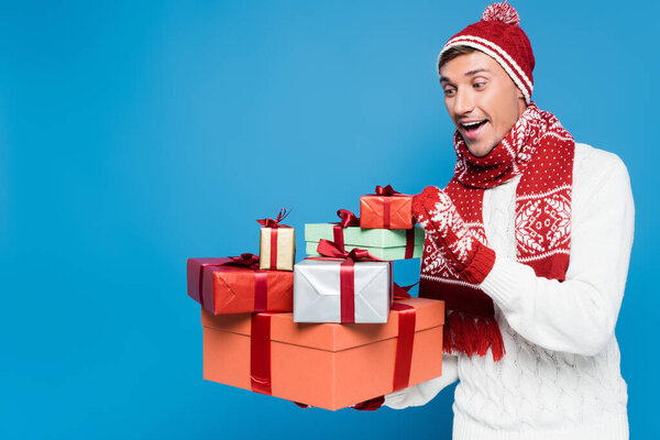 Excited man in knitted hat and mittens holding and looking at bunch of gift boxes isolated on blue