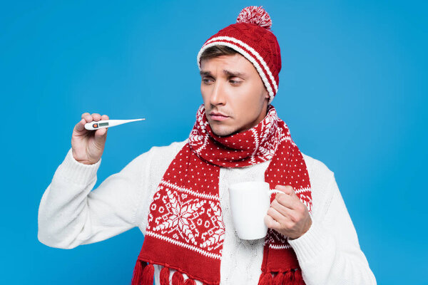 Больной и расстроенный мужчина в вязальной шапочке и с термометром, изолированным на голубом