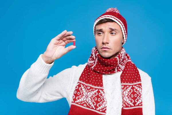 Больной человек в плохом настроении, в теплой одежде, держась и глядя на красную капсулу, изолированную на голубом