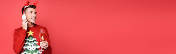Молодой человек в свитере с сосной держит бокал шампанского и разговаривает на смартфоне изолированы на красный, баннер