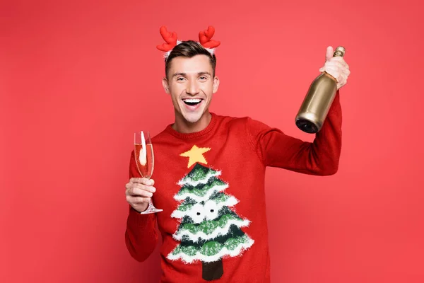 stock image Cheerful man in christmas sweater and headband holding bottle and glass of champagne on red background