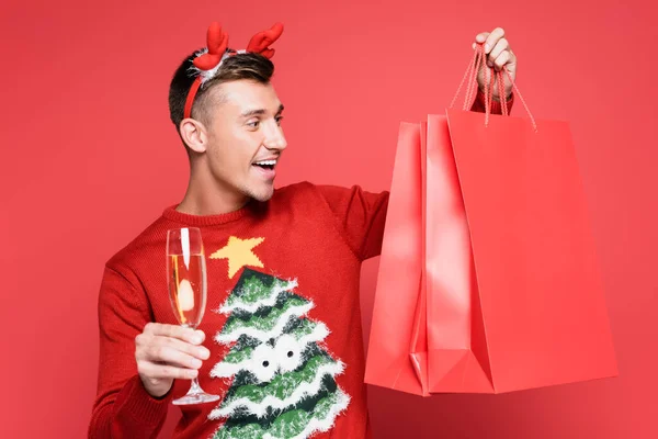 stock image Cheerful man in christmas sweater holding glass of champagne and shopping bags on red background