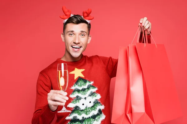 stock image Smiling man in chrismas sweater holding glass of champagne and shopping bags isolated on red