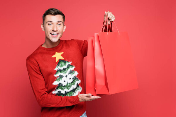 Улыбающийся мужчина в свитере с сосной, держащий сумки с покупками и смотрящий на камеру на красном фоне