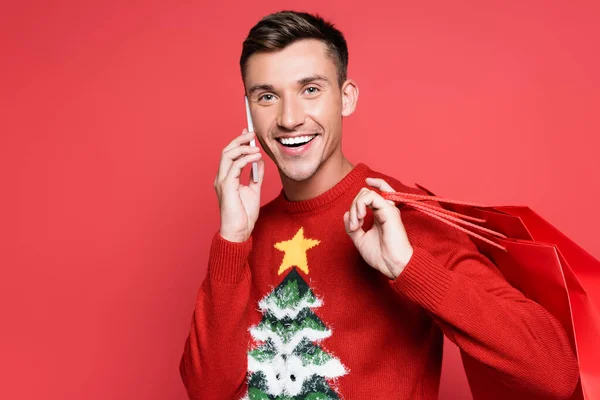 陽気な男でクリスマスセーター話上のスマートフォンや保持ショッピングバッグ孤立した上赤 — ストック写真