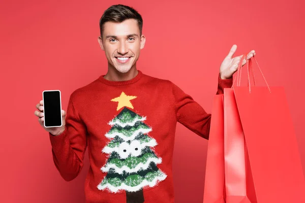 Uśmiechnięty Mężczyzna Swetrze Sosną Trzymający Smartfona Pustym Ekranem Torbami Zakupy — Zdjęcie stockowe