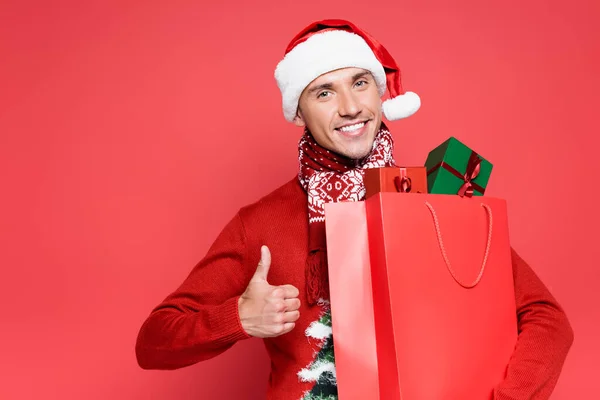 笑顔男でサンタ帽子ショー親指アップしながらショッピングバッグとともにプレゼントオン赤背景 — ストック写真