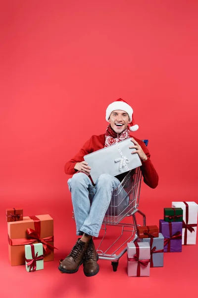 在红色背景的购物车里拿着礼物 戴着圣诞礼帽微笑着看着相机 — 图库照片