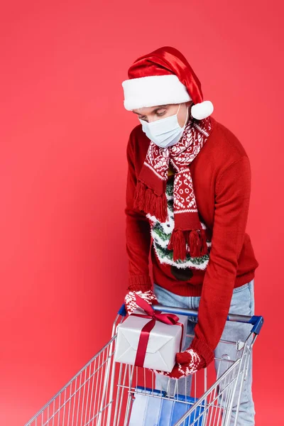 戴着口红礼帽和医疗面具的年轻人在红色背景的购物车旁拿着礼物 — 图库照片