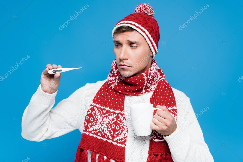 Sick and upset man in knitwear holding cap and showing thermometer isolated on blue