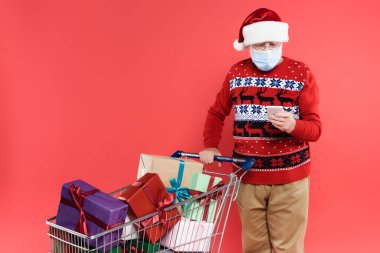 Noel Baba şapkası ve sağlık maskesi takan kıdemli bir adam alışveriş arabasının yanında akıllı telefon kullanıyor.