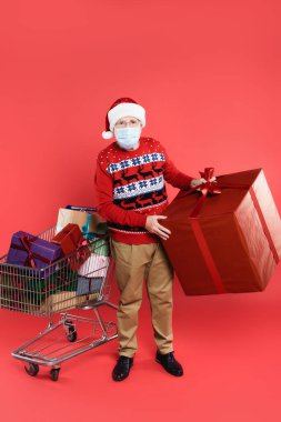 Tıbbi maskeli kıdemli bir adam ve kırmızı arka planda hediyeler olan alışveriş arabasının yanında Noel Baba şapkası tutan bir hediye kutusu.