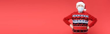 Tıbbi maskeli kıdemli bir adam ve Noel Baba şapkası elleri kalçasında kırmızı pankartta izole bir şekilde duruyor. 