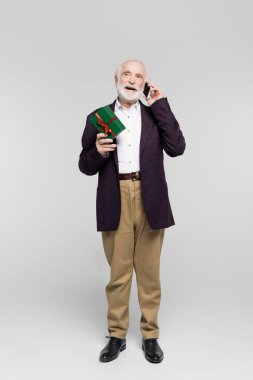 Elinde gri arka planda hediye kutusu tutarken akıllı telefondan konuşan kıdemli bir adam. 