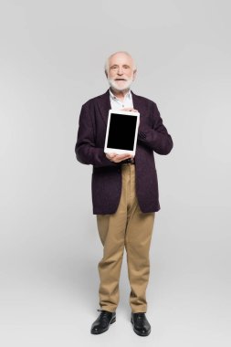 Ceketli yaşlı adam gri arkaplanda boş ekranlı dijital tablet gösteriyor 