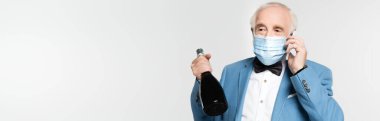 Tıbbi maskeli kıdemli adam akıllı telefondan konuşuyor ve elinde gri pankartta izole edilmiş bir şişe şampanya tutuyor. 