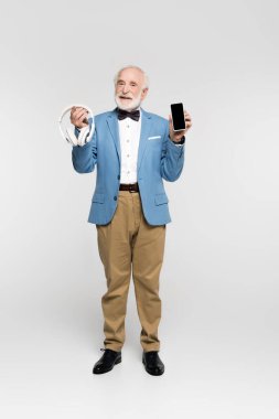 Gri arka planda kablosuz kulaklık ve akıllı telefon tutan gülümseyen yaşlı bir adam. 