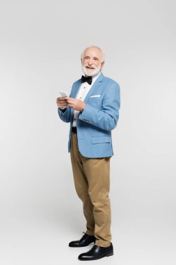 Papyonlu ve ceketli gülümseyen yaşlı adam gri arka planda akıllı telefon tutuyor. 