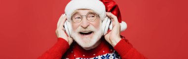 Noel Baba şapkalı gülümseyen yaşlı adam kırmızı pankartta izole edilmiş kulaklıklarla müzik dinliyor.